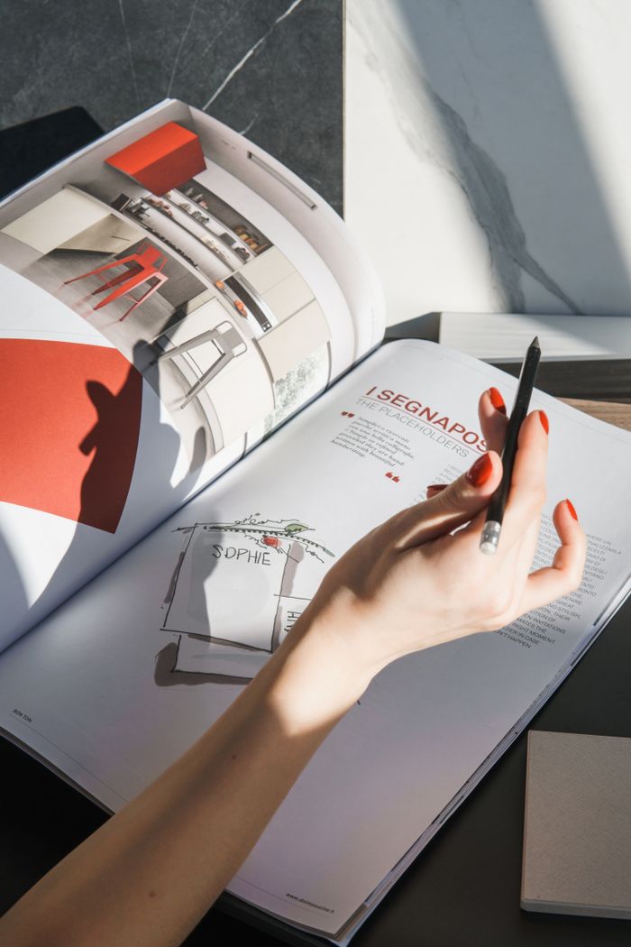 A creative hand holds a pencil over a design magazine, casting a shadow in bright sunlight.